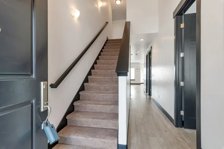Inviting entryway featuring a dark paneled door with brushed nickel hardware, carpeted staircase with dark handrail and trim, and light wood-finish flooring extending into a hallway with recessed lighting and dark-trimmed doorways