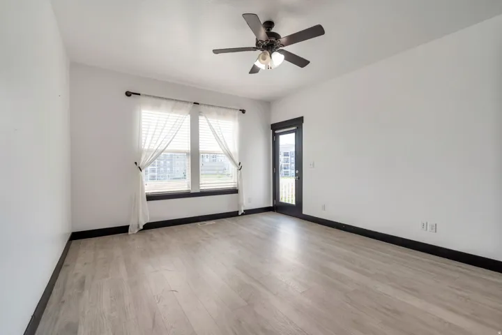Spacious room featuring wood-finish flooring, a ceiling fan with integrated lighting, and a large window with dark trim