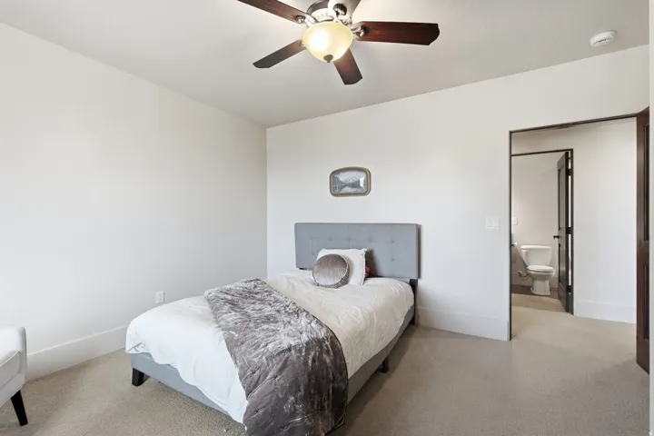 Carpeted room featuring a ceiling fan with integrated lighting, white baseboards, and a dark-framed doorway leading to an adjoining bathroom with a toilet and dark flooring
