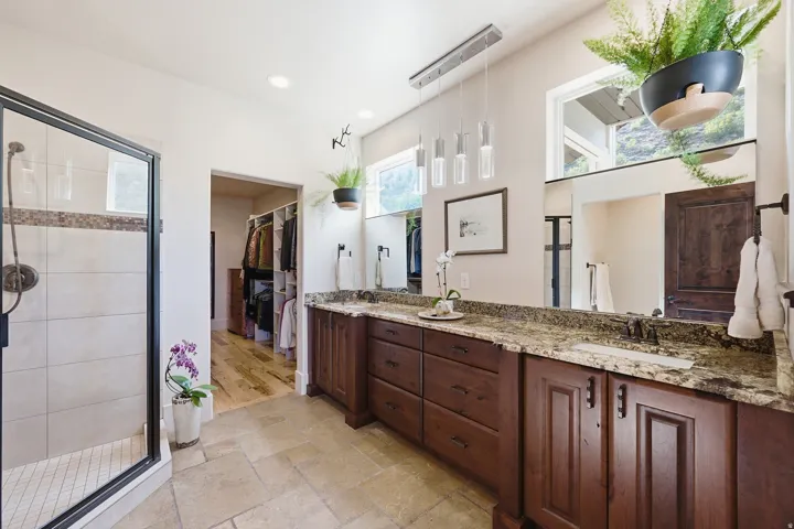 Spacious bathroom featuring a dual vanity with a granite countertop, rich wood cabinetry, and a walk-in shower with tile surround