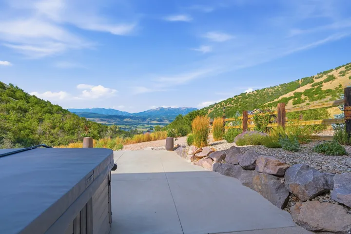Paved patio area featuring a hot tub, rock retaining wall, landscaped garden beds, and a wooden fence