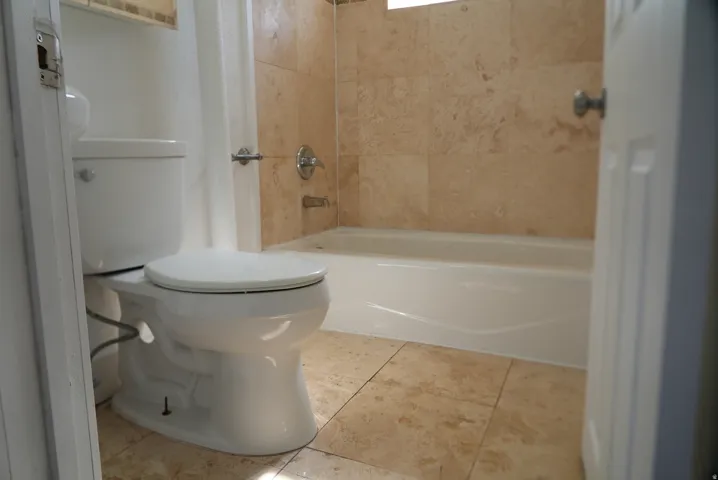Contemporary bathroom featuring a white toilet, tile flooring, a white bathtub, and floor-to-ceiling tiled walls in the bathing area
