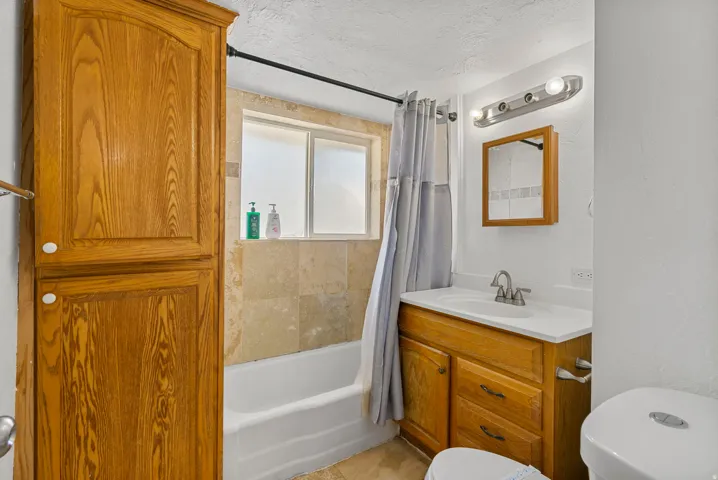 Bathroom featuring a built-in wood-finish storage cabinet, a white bathtub with stone-finish tile surround, a window with privacy glass, a vanity with wood-finish cabinetry, and a mirrored medicine cabinet