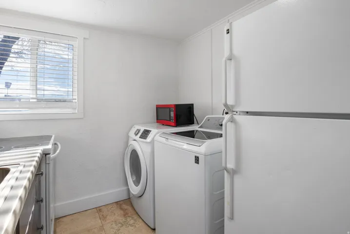 Utility area featuring a white top-freezer refrigerator, front-load washing machine, top-load washing machine, and a window with horizontal blinds