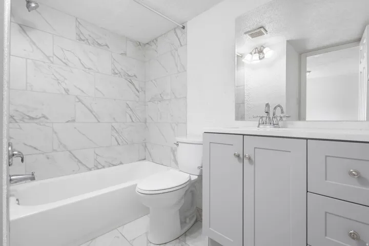 Bathroom featuring marble-patterned tile, a white bathtub, a toilet, a light gray vanity with chrome hardware, and a mirrored medicine cabinet