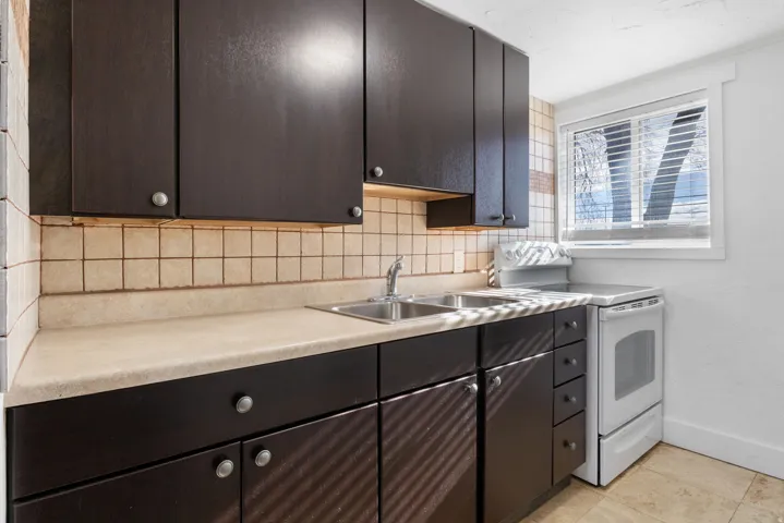 Kitchen featuring dark wood-finish cabinetry, a light-toned countertop, a tile backsplash, and a window with blinds
