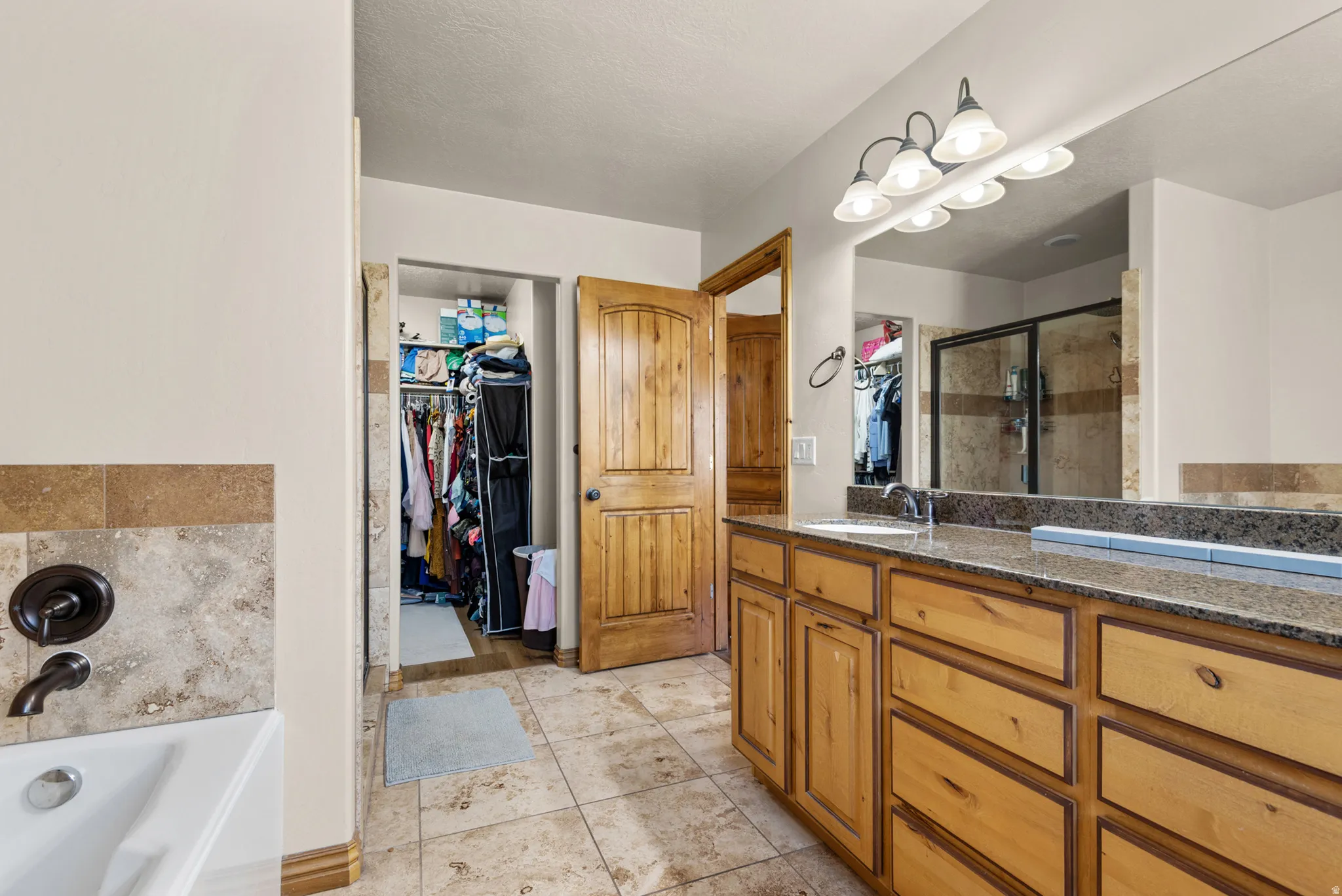Tiled bathroom featuring a dual vanity with light wood cabinetry and granite-look countertops, a built-in tub with dark bronze fixtures, and a shower enclosure with glass doors
