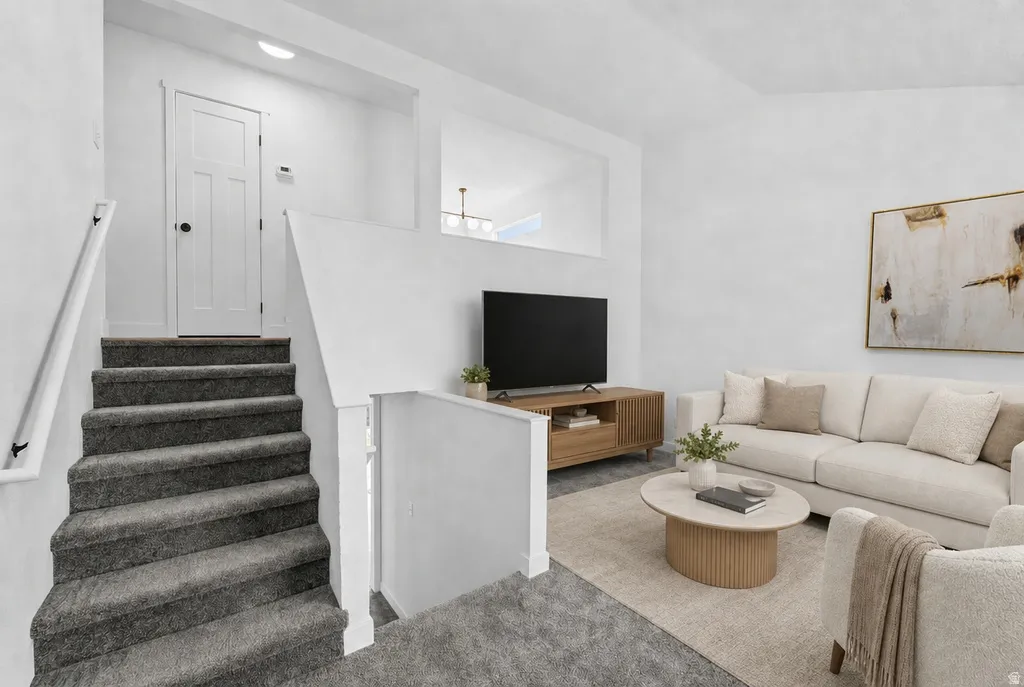 Finished basement recreation area featuring a gray carpeted staircase with white railings, white drywall partitions, recessed ceiling lighting, a wood-finish media console with flat-screen television, neutral upholstered seating, a round ottoman with wove