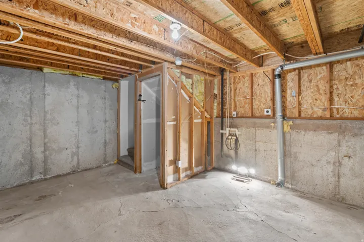Unfinished basement space featuring exposed wood joists, concrete walls, a concrete floor, and visible utility conduits