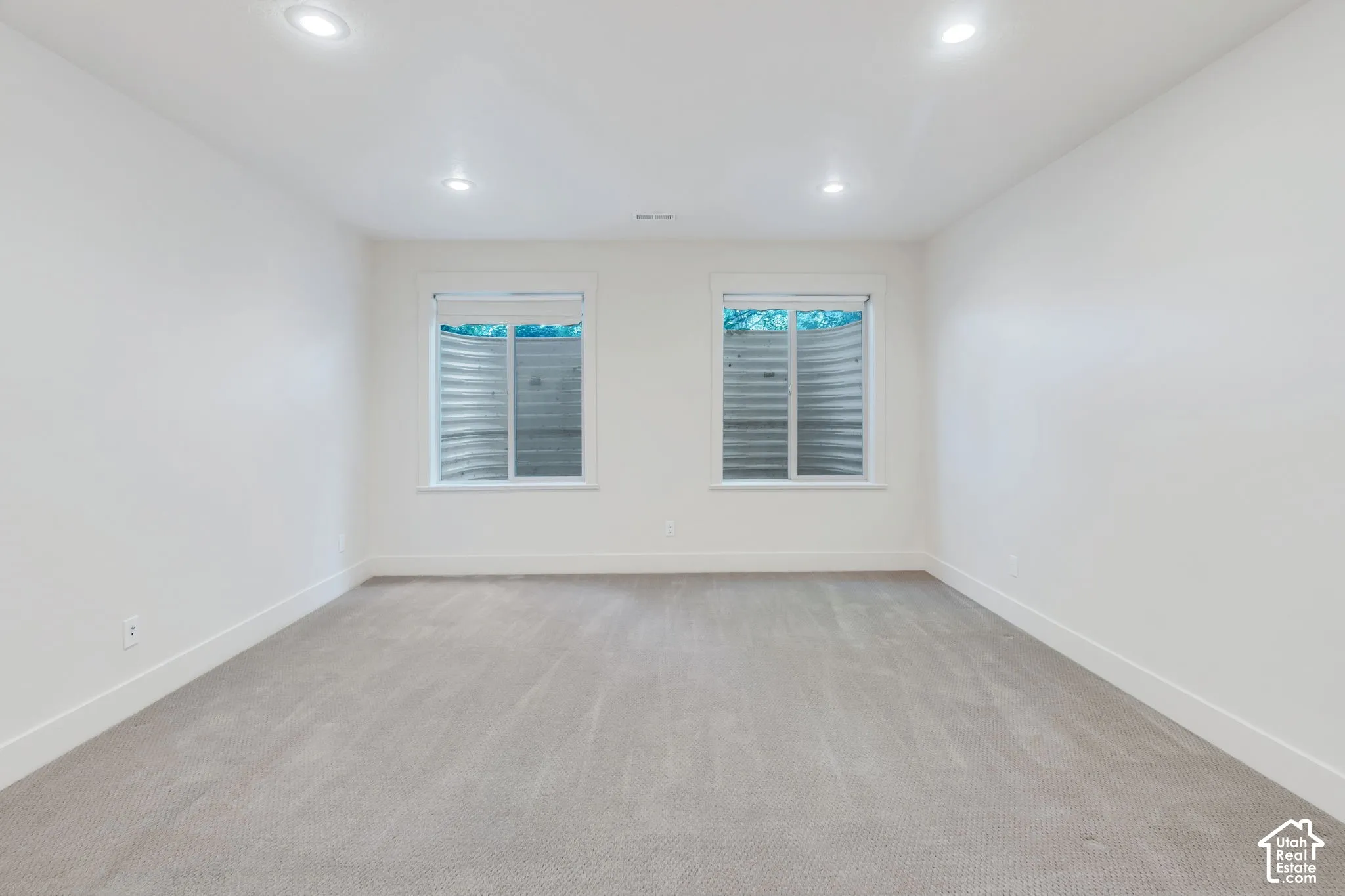 Spacious room featuring light gray carpeting, two egress windows with visible window wells, white walls, white baseboards, and recessed ceiling lighting