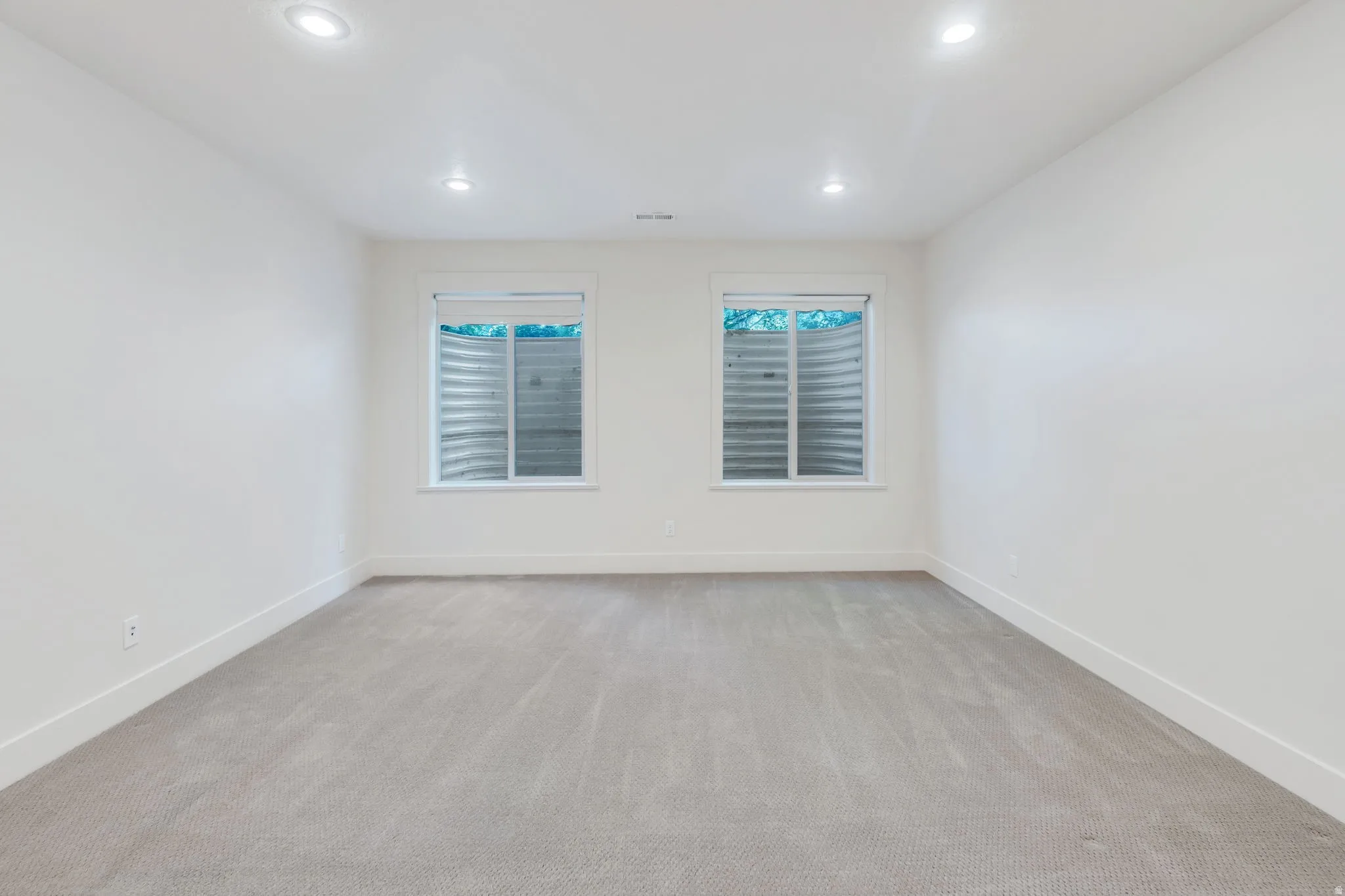 Spacious room featuring light gray carpeting, two egress windows with visible window wells, white walls, white baseboards, and recessed ceiling lighting