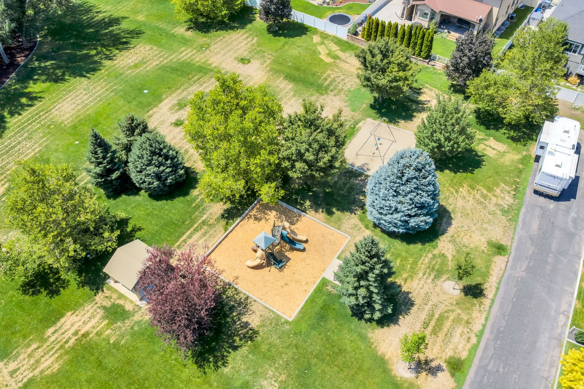 Expansive green space featuring a playground with wood chip surfacing, a swing set, mature trees, and a small outbuilding with a brown shingled roof