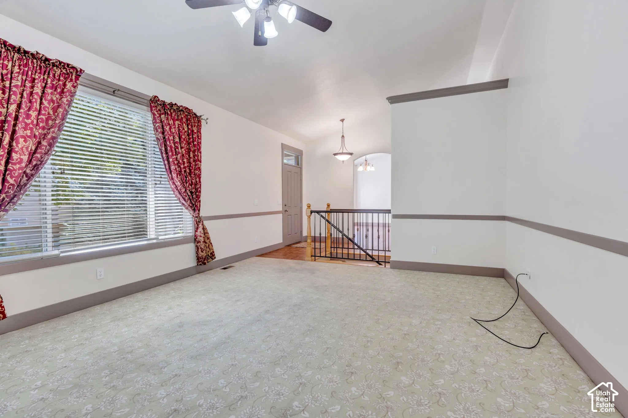 Spacious upper-level room featuring neutral carpet flooring, a ceiling fan with integrated lighting, large window with red patterned curtains and horizontal blinds, chair rail molding detail, and a safety gate