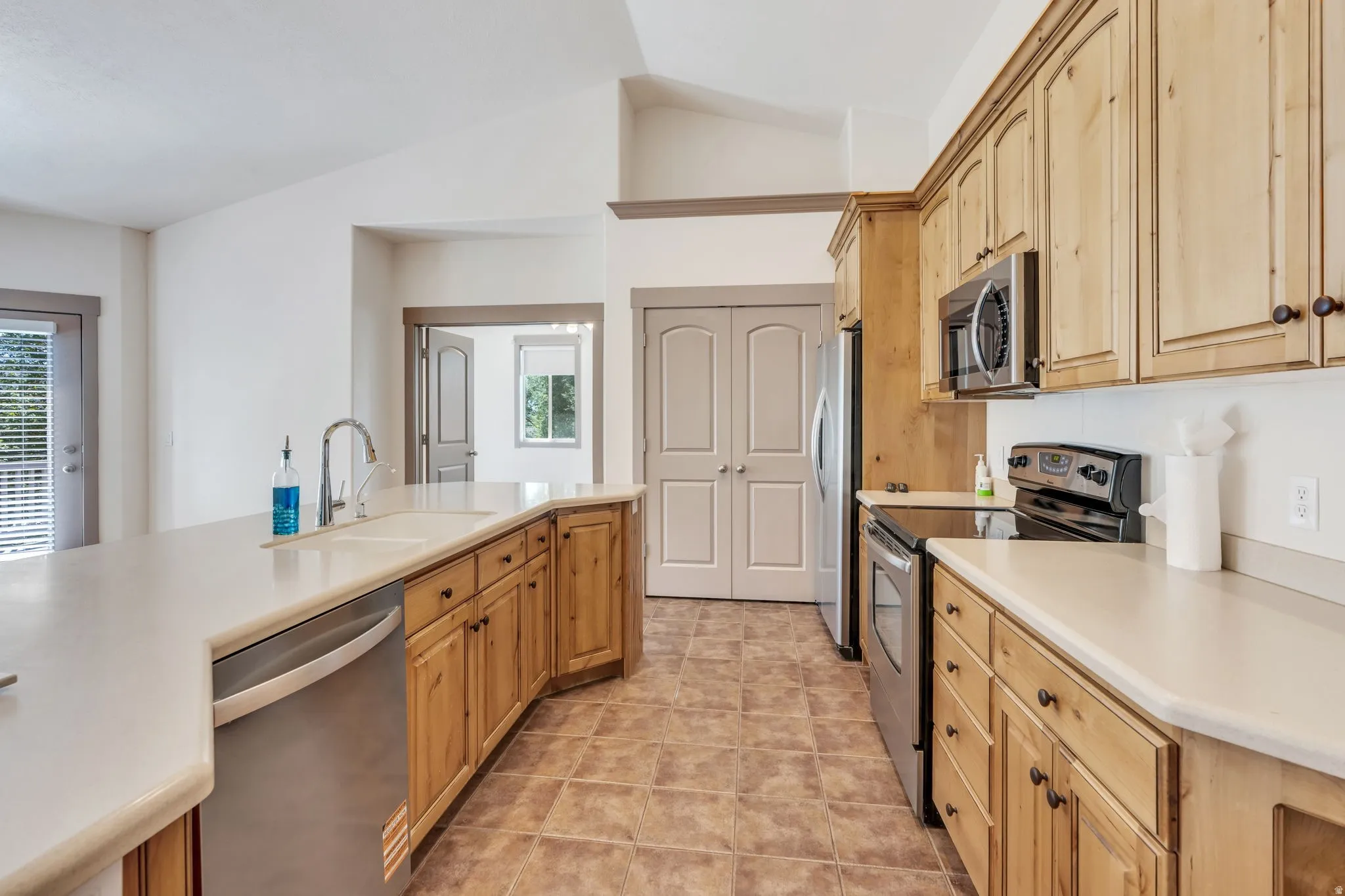 Kitchen featuring wood-finish cabinetry, stainless steel appliances, light-toned countertops, tile flooring, and vaulted ceilings