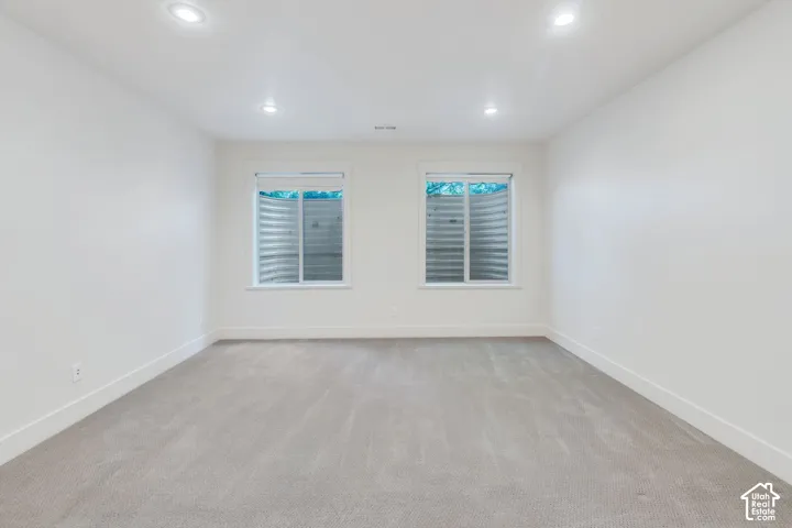 Spacious room featuring light gray carpeting, two egress windows with visible window wells, white walls, white baseboards, and recessed ceiling lighting