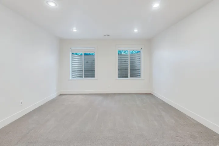 Spacious room featuring light gray carpeting, two egress windows with visible window wells, white walls, white baseboards, and recessed ceiling lighting