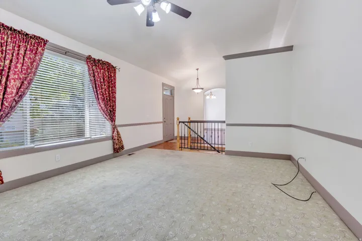 Spacious upper-level room featuring neutral carpet flooring, a ceiling fan with integrated lighting, large window with red patterned curtains and horizontal blinds, chair rail molding detail, and a safety gate
