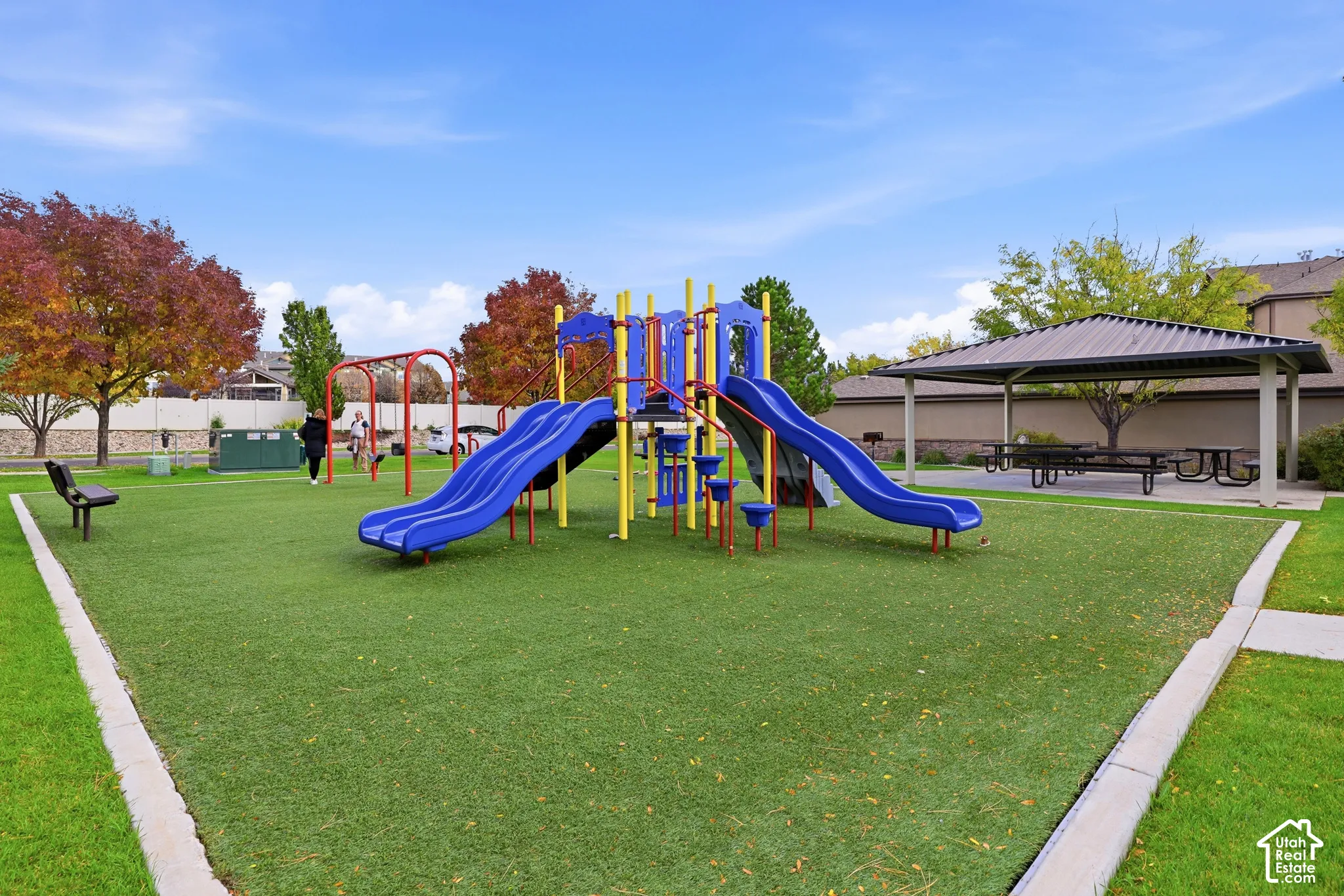 Outdoor recreation area featuring a multi-component play structure, covered pavilion, synthetic turf surfacing, and peripheral park bench seating