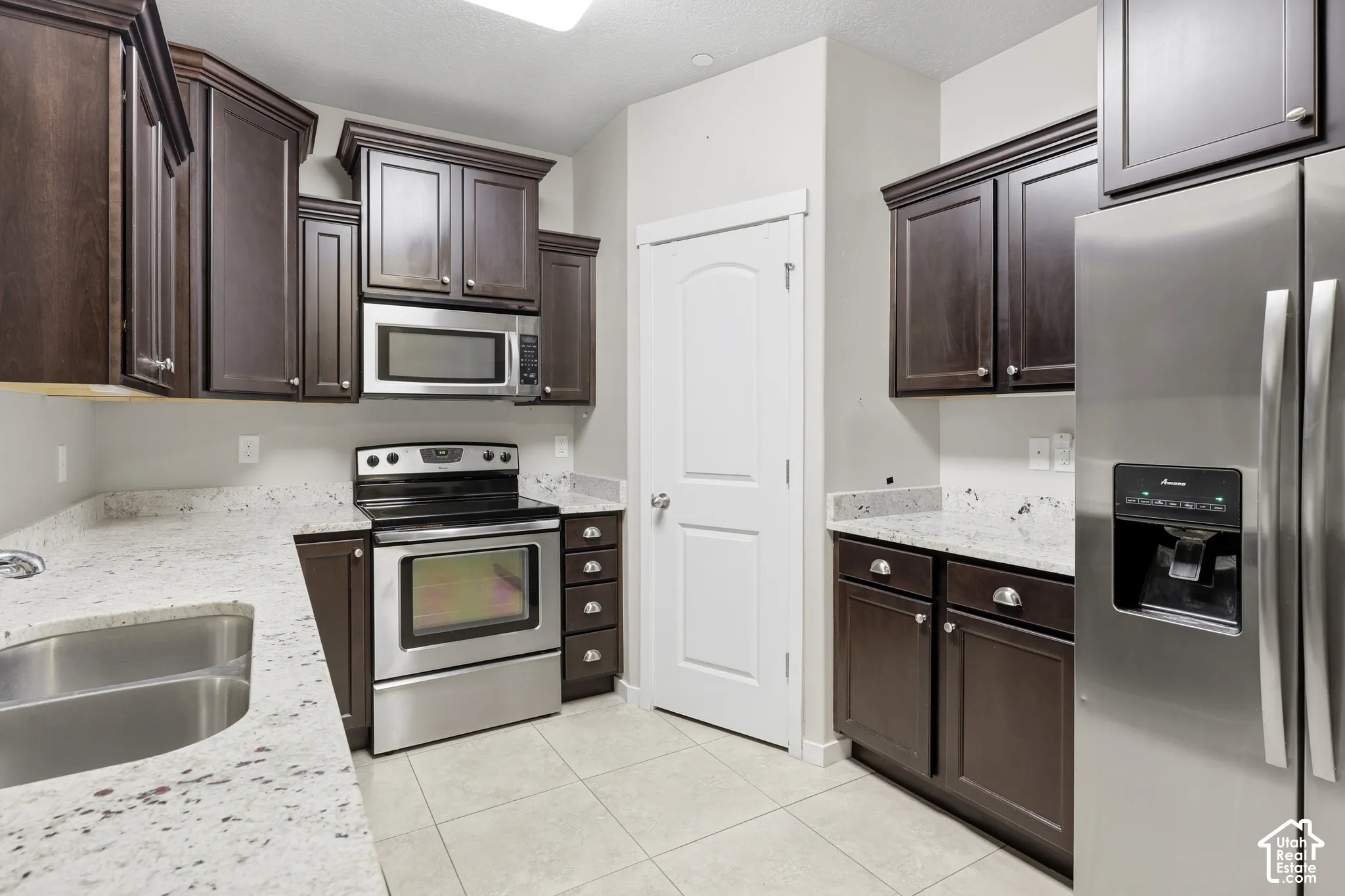 Kitchen featuring dark-finish cabinetry, light-toned stone-patterned countertops, stainless steel appliances, and tile flooring