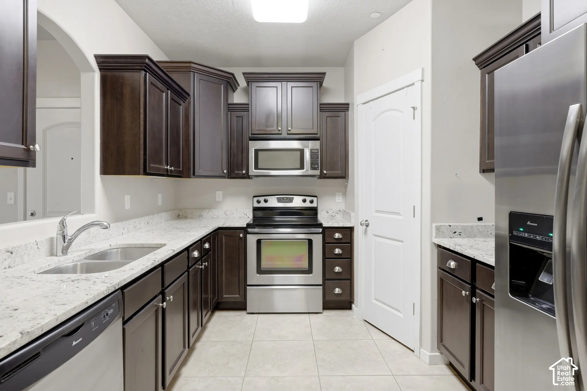 Kitchen featuring dark wood-finish cabinetry, stone-surface countertops, stainless steel appliances, a double-basin undermount sink, and tiled flooring