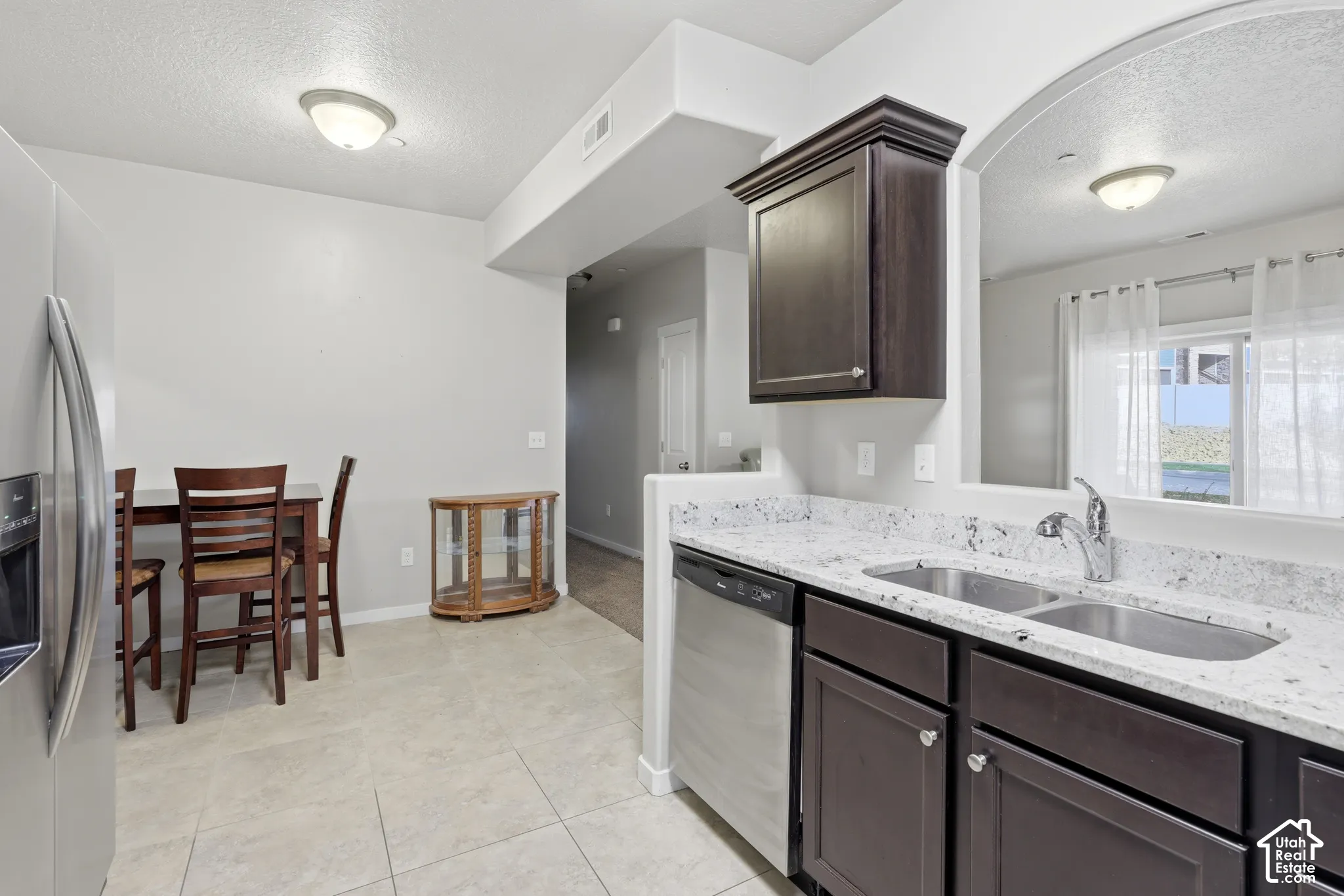 Kitchen featuring dark-finish cabinetry, light-toned stone-patterned countertops, stainless steel appliances, dual-basin sink, and tile flooring
