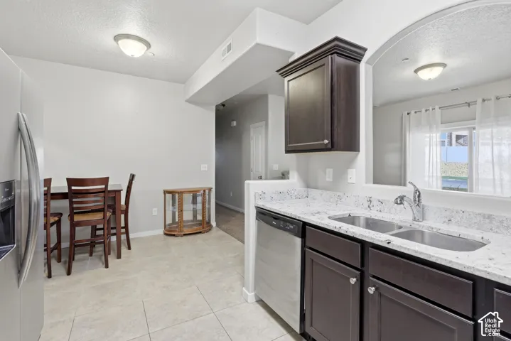 Kitchen featuring dark-finish cabinetry, light-toned stone-patterned countertops, stainless steel appliances, dual-basin sink, and tile flooring