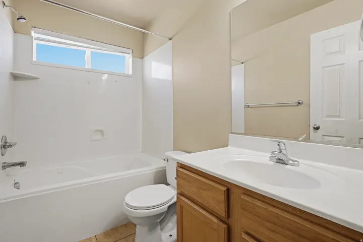 Bathroom featuring a white bathtub with an integrated shower, a vanity with a white countertop and natural wood-finish cabinetry, a toilet, and a window above the tub