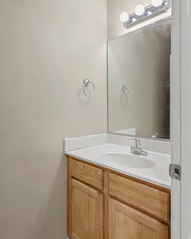Bathroom vanity with a white countertop and integrated sink, chrome faucet, wood-finish cabinetry, frameless wall mirror, and a three-bulb vanity light fixture