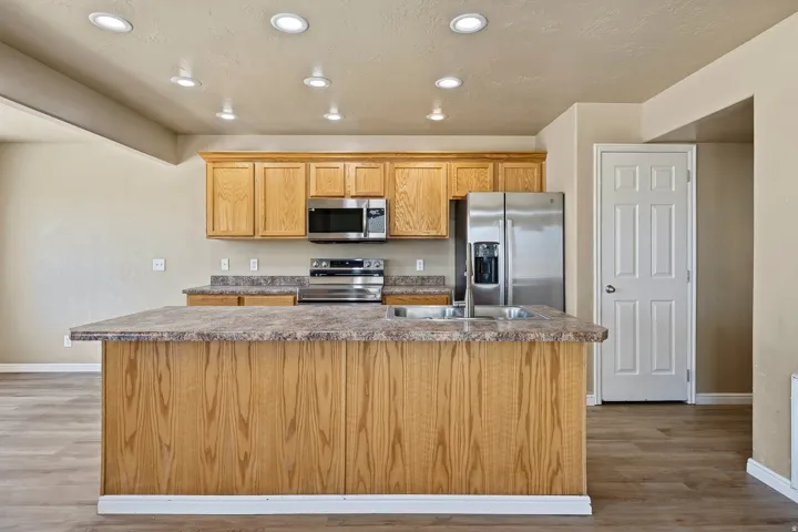 Kitchen featuring a central island with an integrated sink, natural wood cabinetry, stainless steel appliances, recessed ceiling lighting, and wood-finish flooring