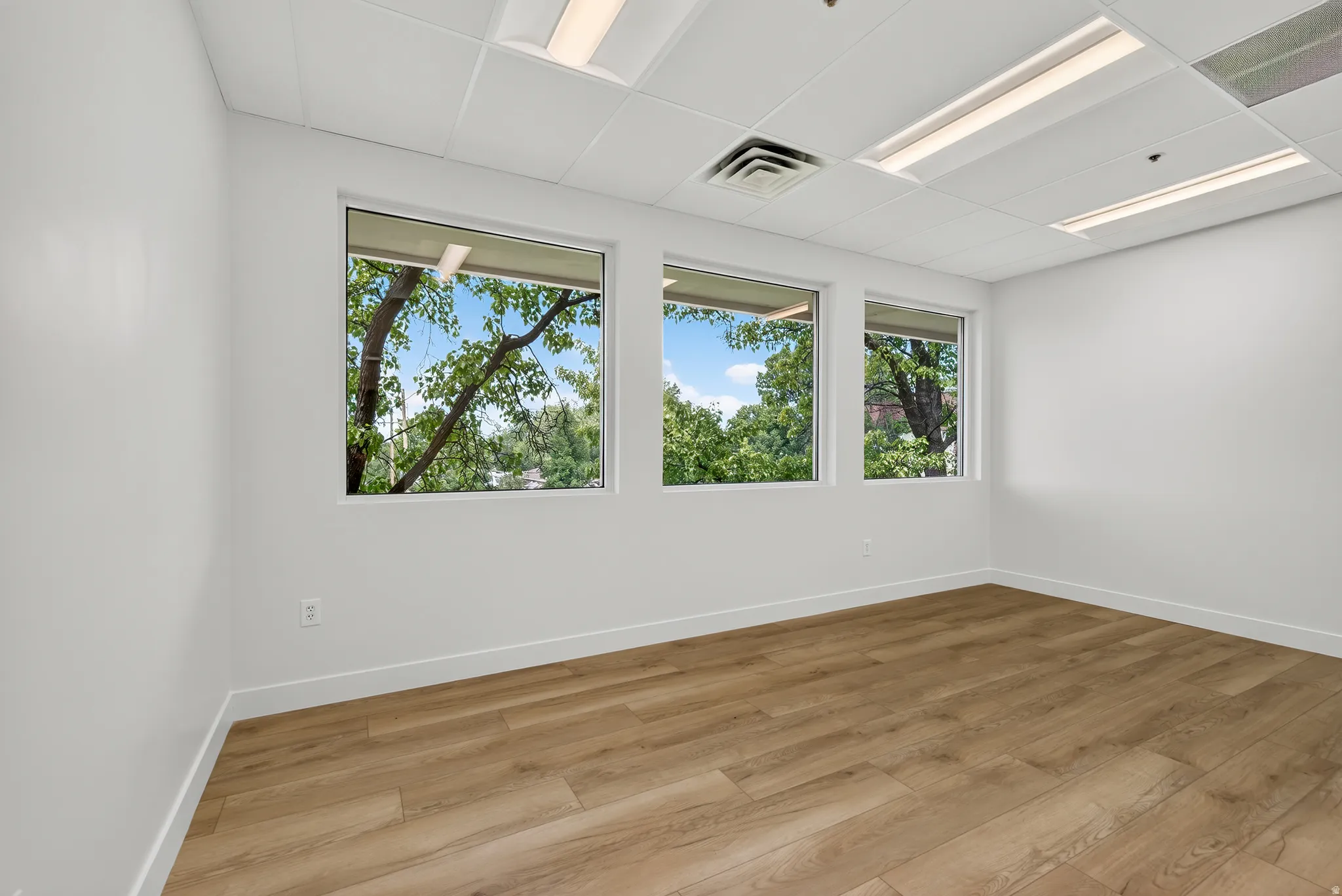Bright interior space featuring three large windows, wood-finish flooring, and a drop ceiling with integrated lighting