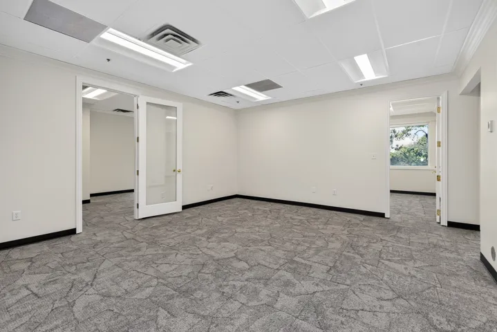 Spacious interior with textured grey carpeting, white walls, drop ceiling with integrated lighting, multiple doorways, and one window offering natural light