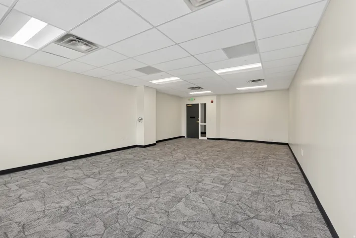 Open-concept interior featuring light gray patterned carpet, drop ceiling with recessed lighting, off-white walls, black baseboards, and a dark entry door with a glass panel