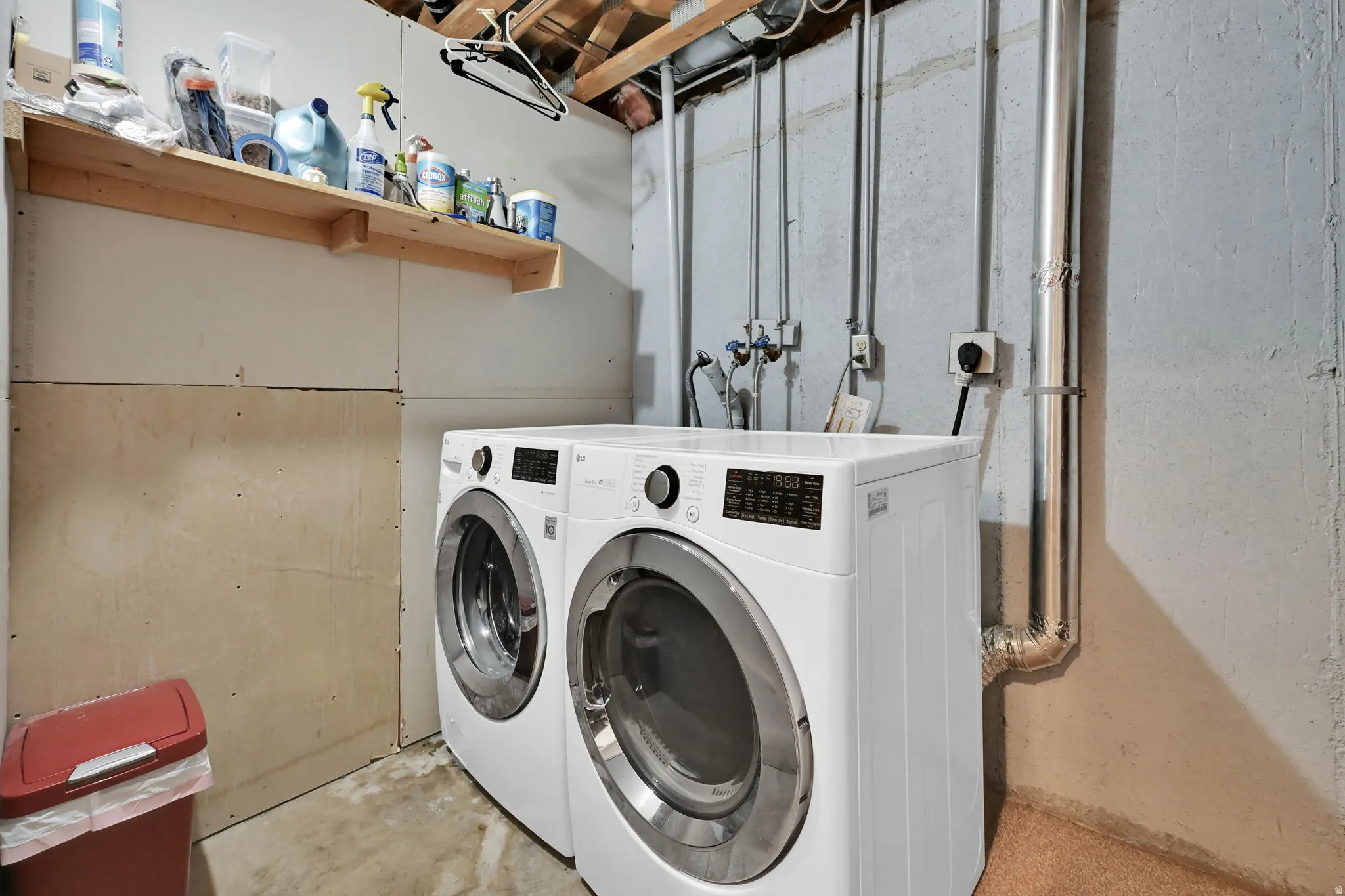 Laundry area featuring washer and dryer hookups, exposed ceiling joists, utility piping, and a wall-mounted shelving unit