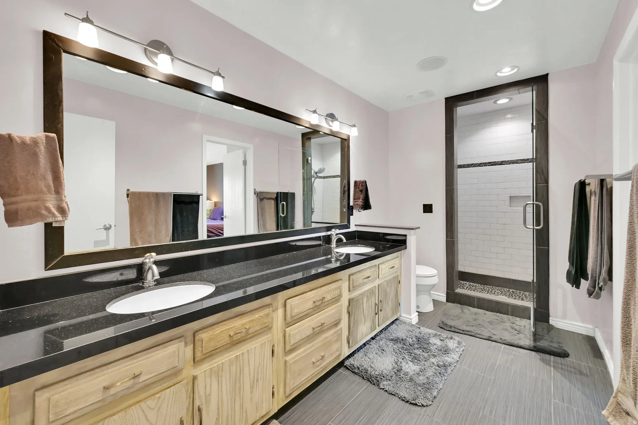 Dual vanity with dark countertop and undermount sinks, expansive framed mirror, dedicated shower with glass enclosure, white subway tile shower surround, and gray tile flooring