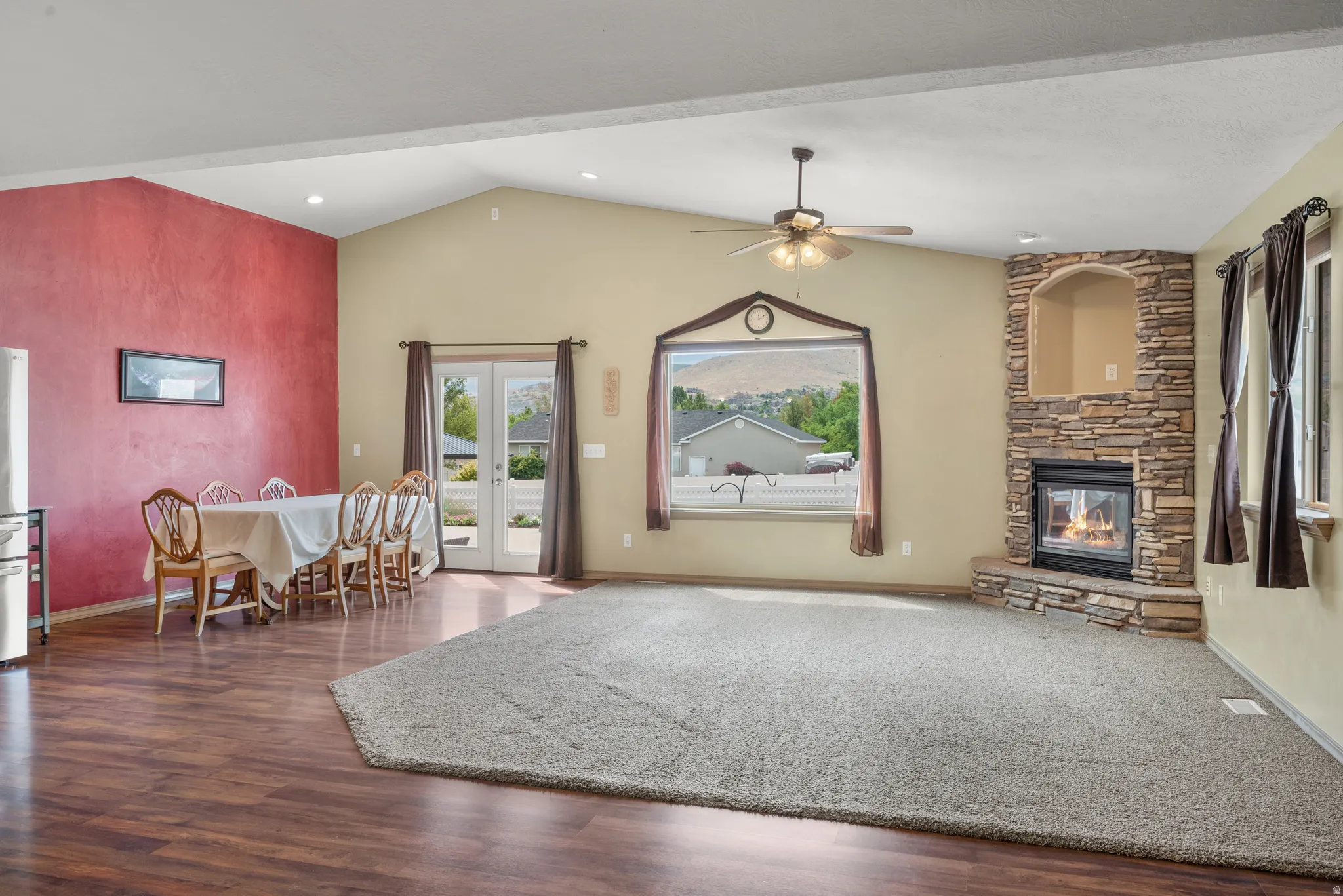 Spacious open-concept living area featuring warm wood-finish flooring, a stone fireplace with gas insert, vaulted ceiling with ceiling fan, and abundant natural light from multiple windows and glass doors