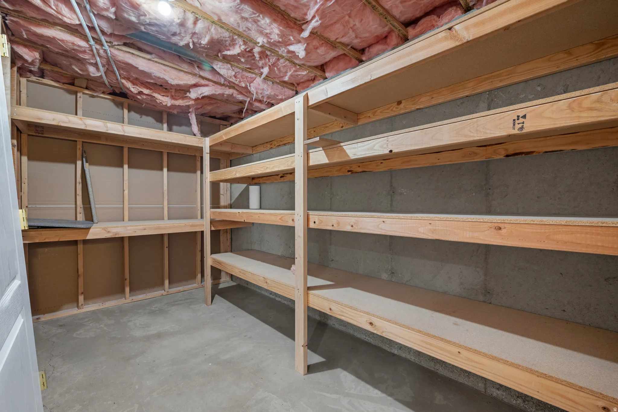 Utility room featuring extensive built-in wooden shelving with multiple tiers, concrete flooring, exposed wood beam ceiling structure, and white drywall paneling on the left wall for additional storage and organization