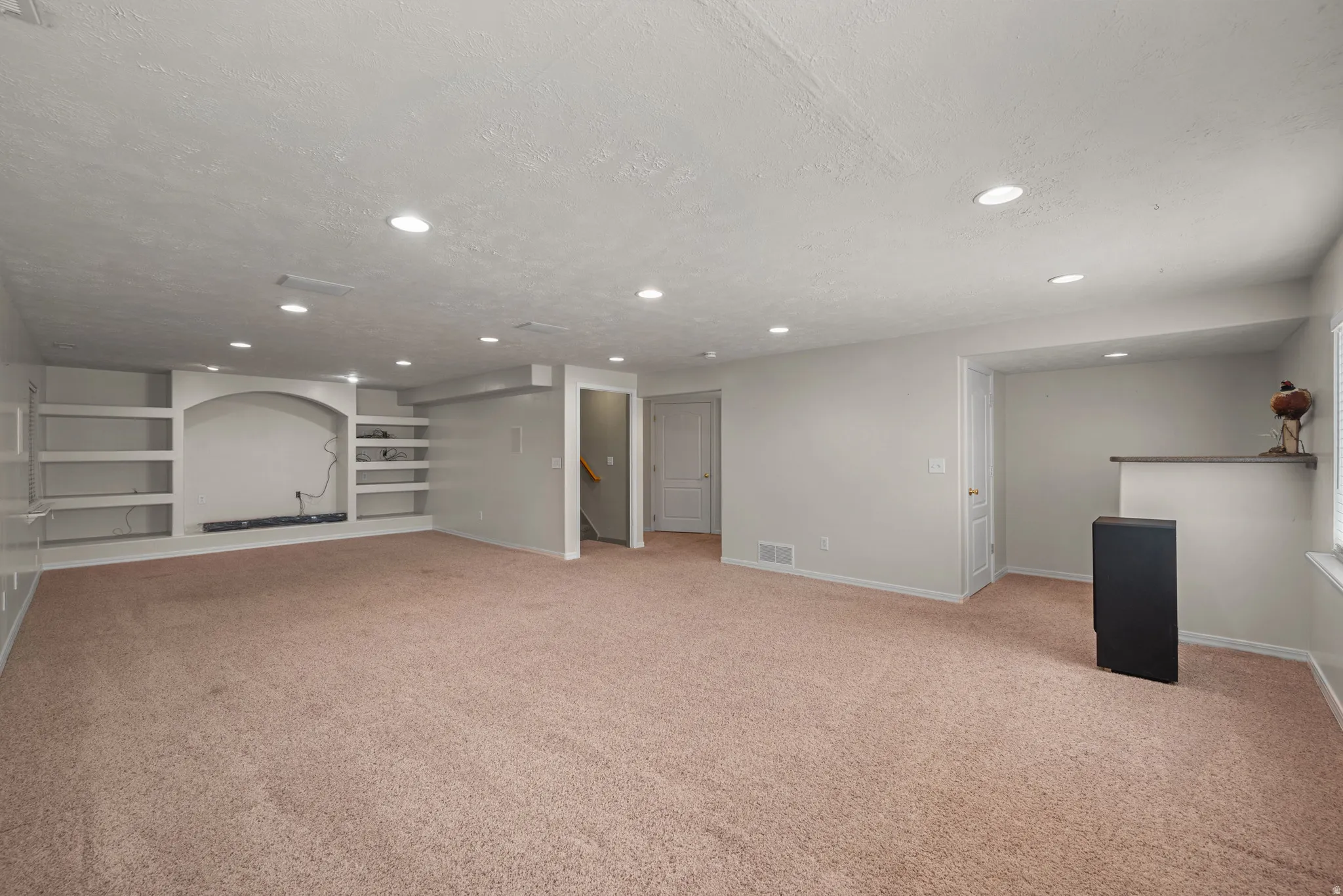 Spacious basement recreation area featuring neutral carpeting, recessed ceiling lighting, built-in shelving with arched alcove detail, and multiple access points