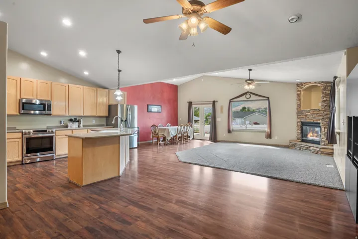 Open-concept living space featuring warm wood-finish flooring throughout, custom cabinetry with stainless steel appliances, a large center island with seating, pendant lighting, ceiling fans with integrated fixtures, and a fireplace with built-in surround