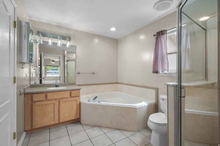 Spa-inspired primary bathroom featuring a soaking tub with tile surround, wood-finish vanity with dual sink configuration, recessed lighting, ceramic tile flooring, and a frameless glass-enclosed shower enclosure with tile walls