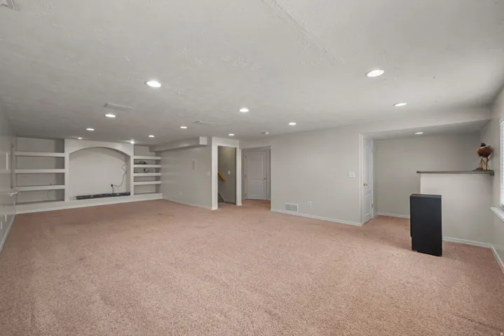 Spacious basement recreation area featuring neutral carpeting, recessed ceiling lighting, built-in shelving with arched alcove detail, and multiple access points