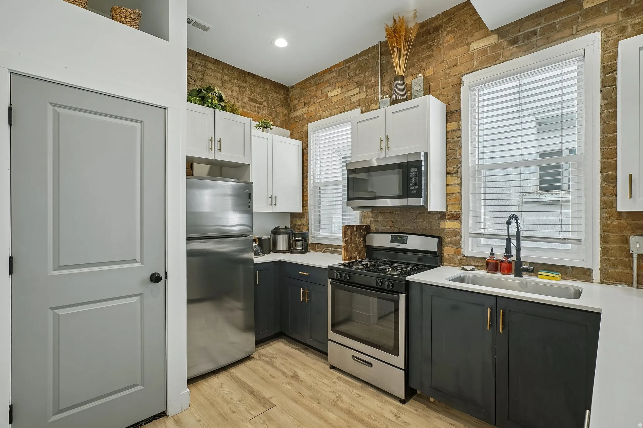 Exposed brick wall, stainless steel appliances, wood-finish flooring, two-tone cabinetry, and a stainless steel sink with a matte black faucet