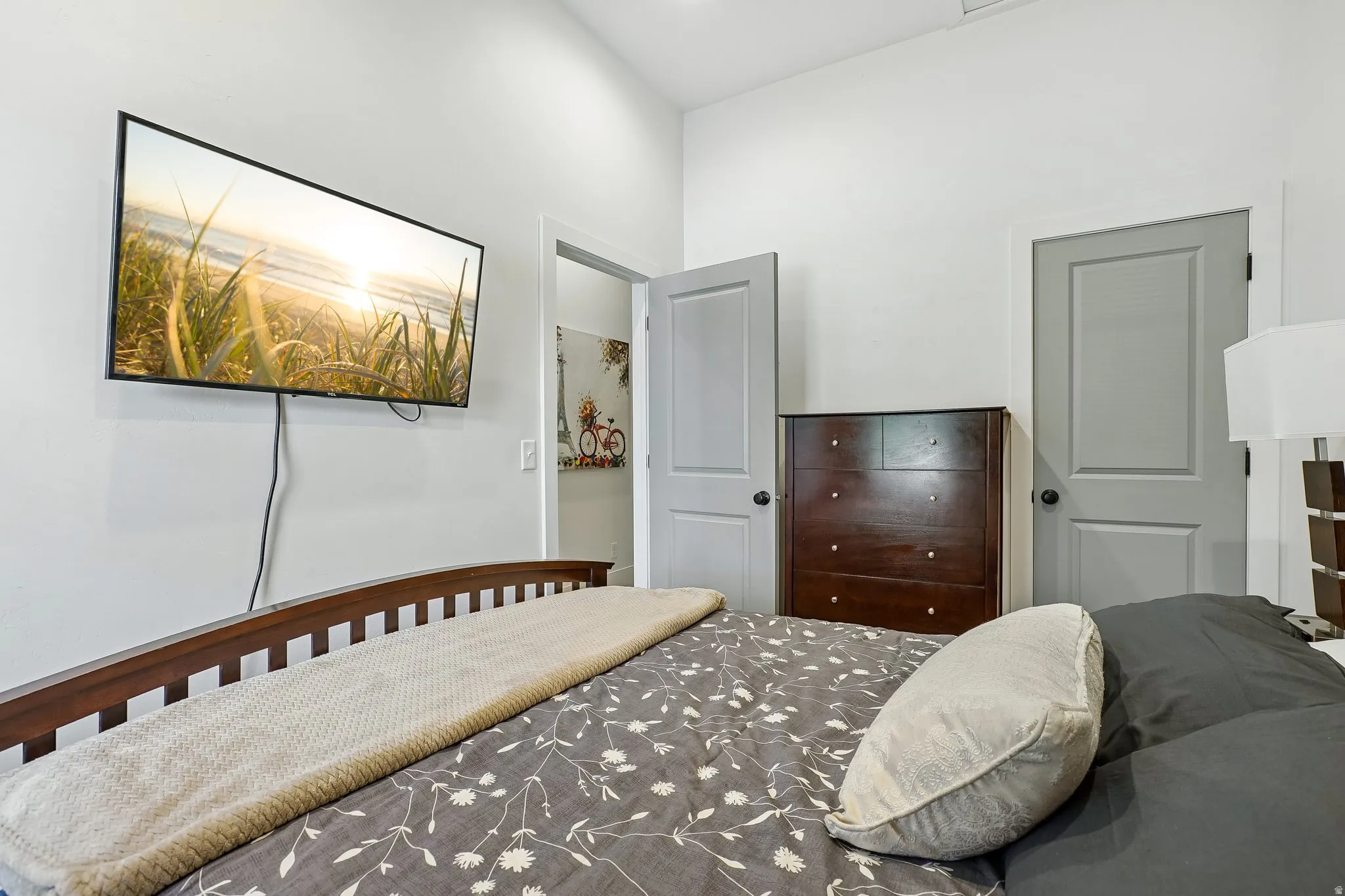 Bright secondary bedroom featuring warm wood-finish flooring, a wood-frame bed, dark wood dresser with multiple drawers, and dual gray-painted doors providing access to adjoining spaces