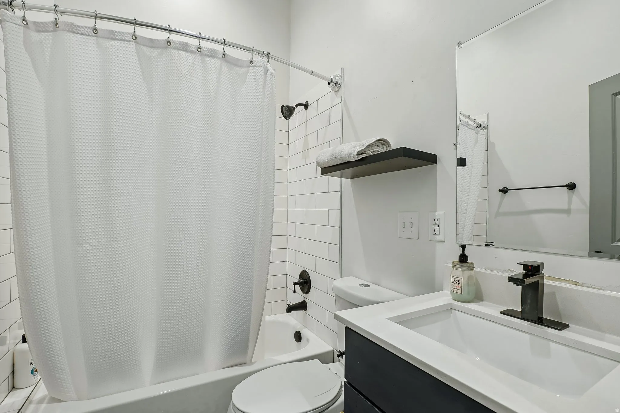 Bathtub with subway tile surround, dark bronze shower fixtures, floating vanity with white countertop, integrated sink, and wall-mounted mirror