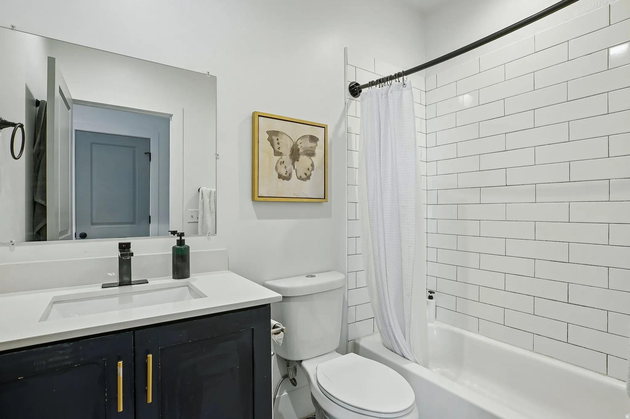 Contemporary bathroom featuring a black vanity with brass hardware and white countertop, white subway tile surround, framed mirror with integrated lighting, and a white subway tile tub/shower enclosure with chrome fixtures and white curtain rod