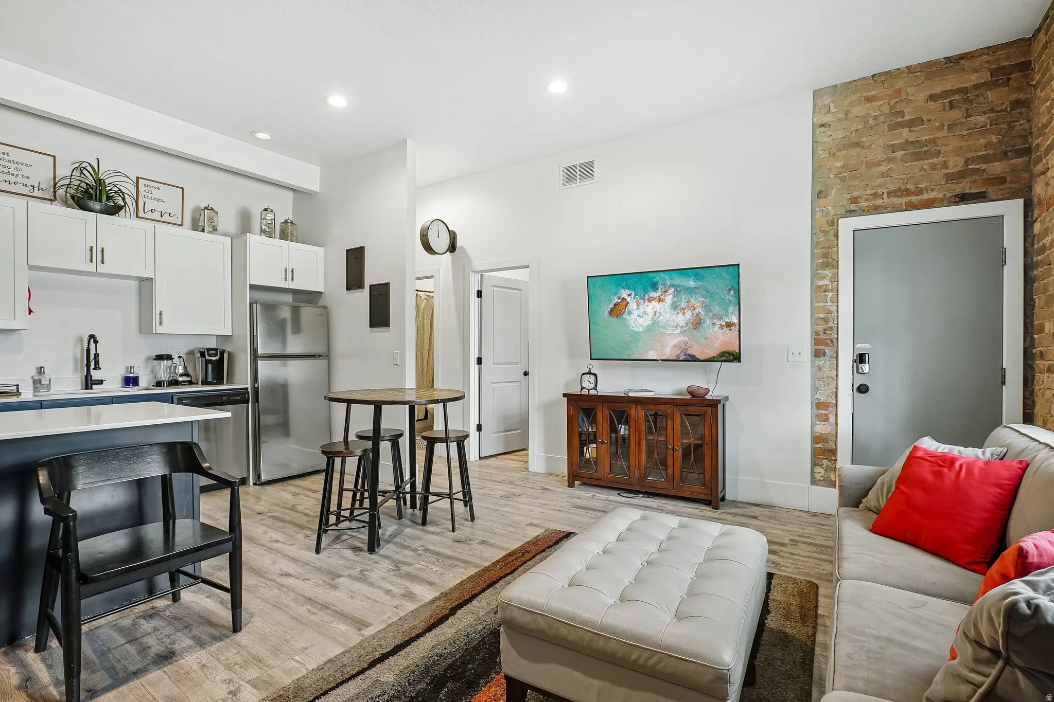 Open-concept living area featuring wood-finish flooring, stainless steel appliances, white cabinetry with hardware accents, exposed brick accent wall, and a wall-mounted flat-screen television above a wooden media console
