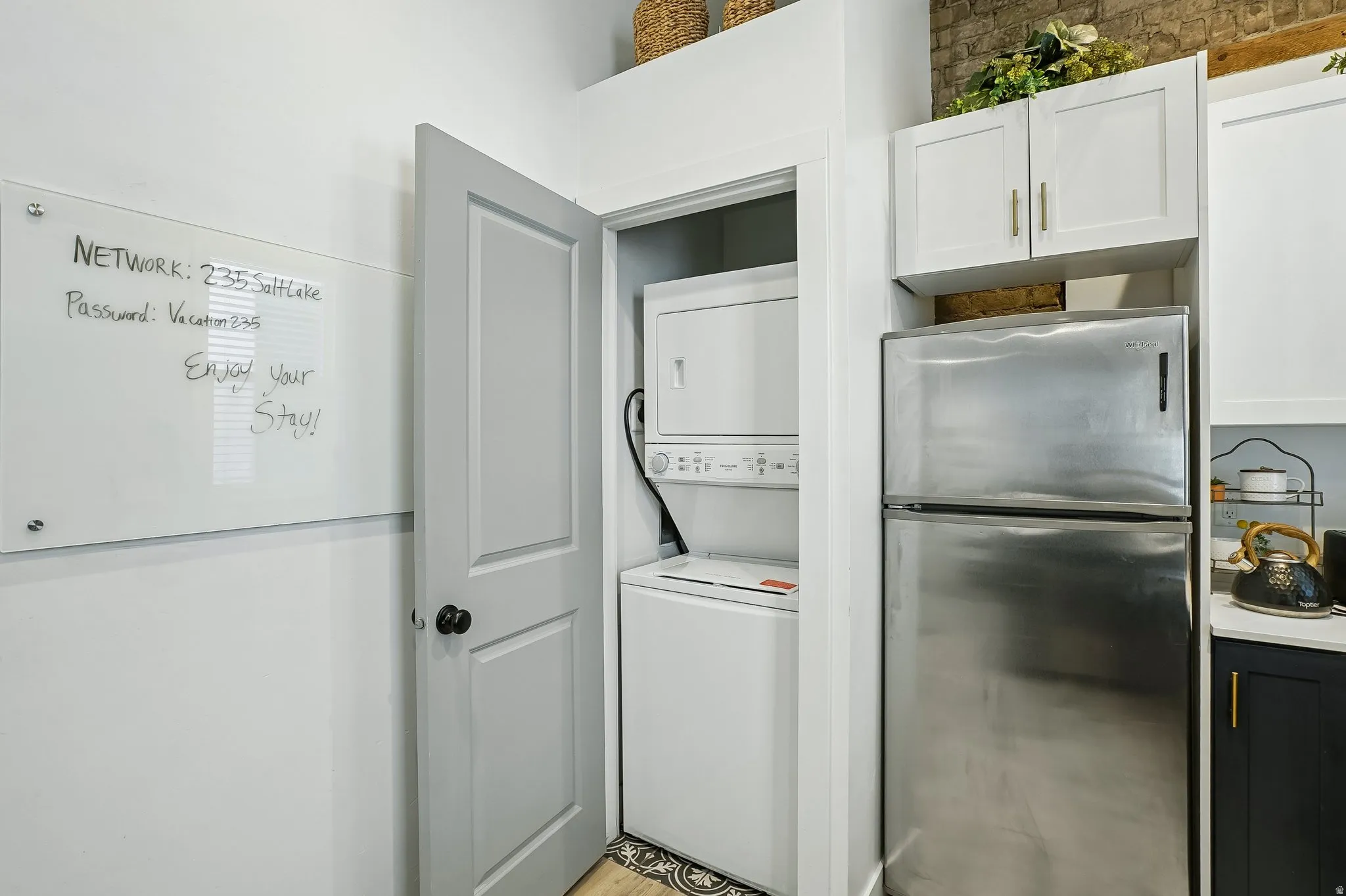 Integrated stackable washer and dryer, stainless steel refrigerator, white upper cabinetry, and exposed brick detailing