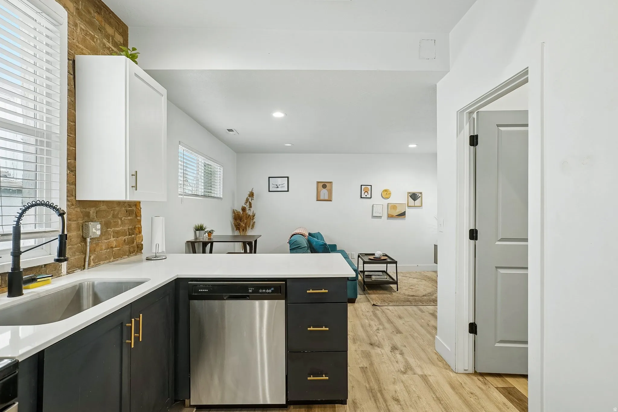 Modern kitchen featuring stainless steel appliances, black cabinetry with brass hardware, white quartz countertops, exposed brick backsplash, and wood-finish flooring throughout the open layout