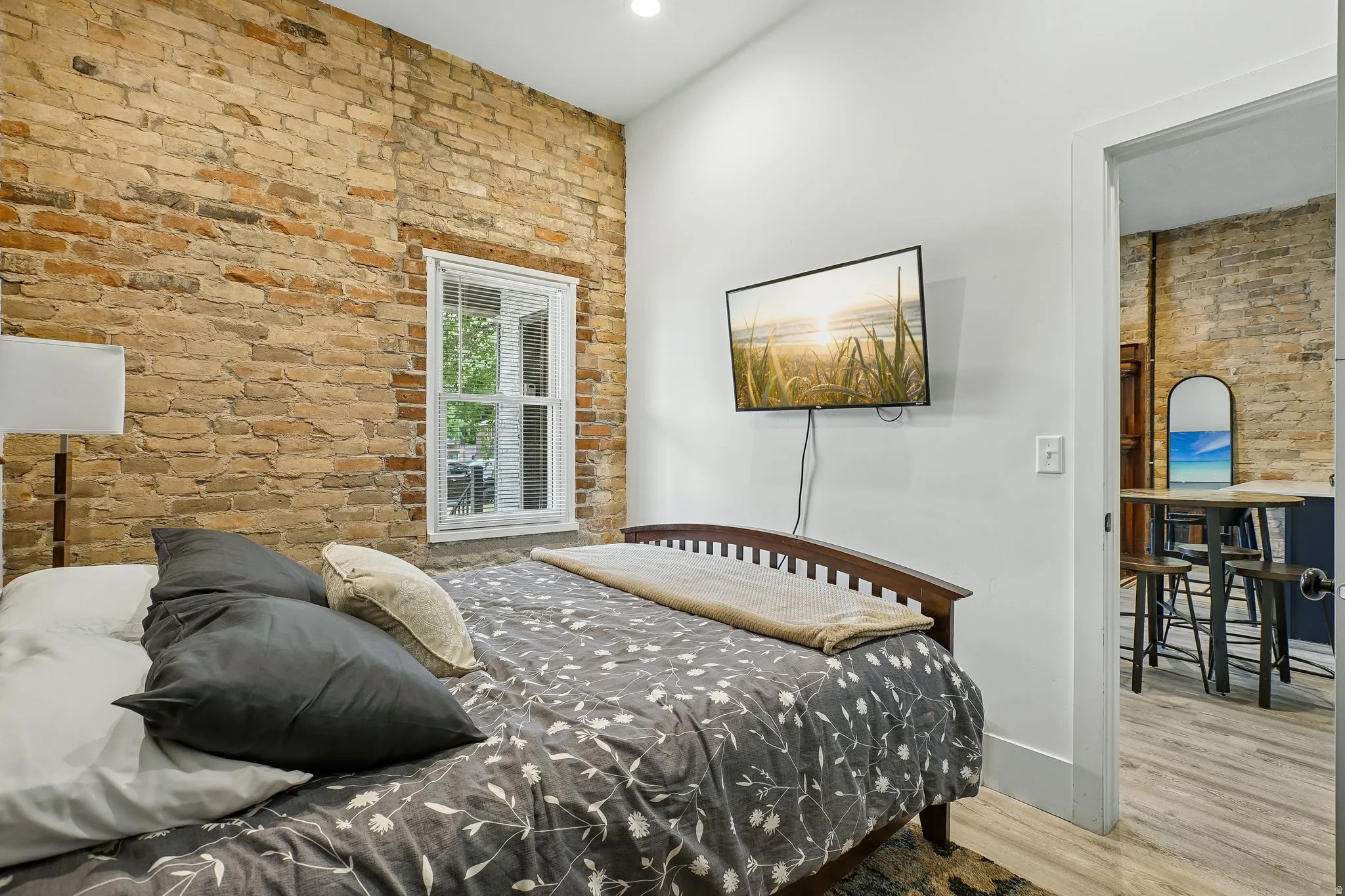 Bedroom featuring exposed brick accent wall, wood-finish flooring, and abundant natural light from large window