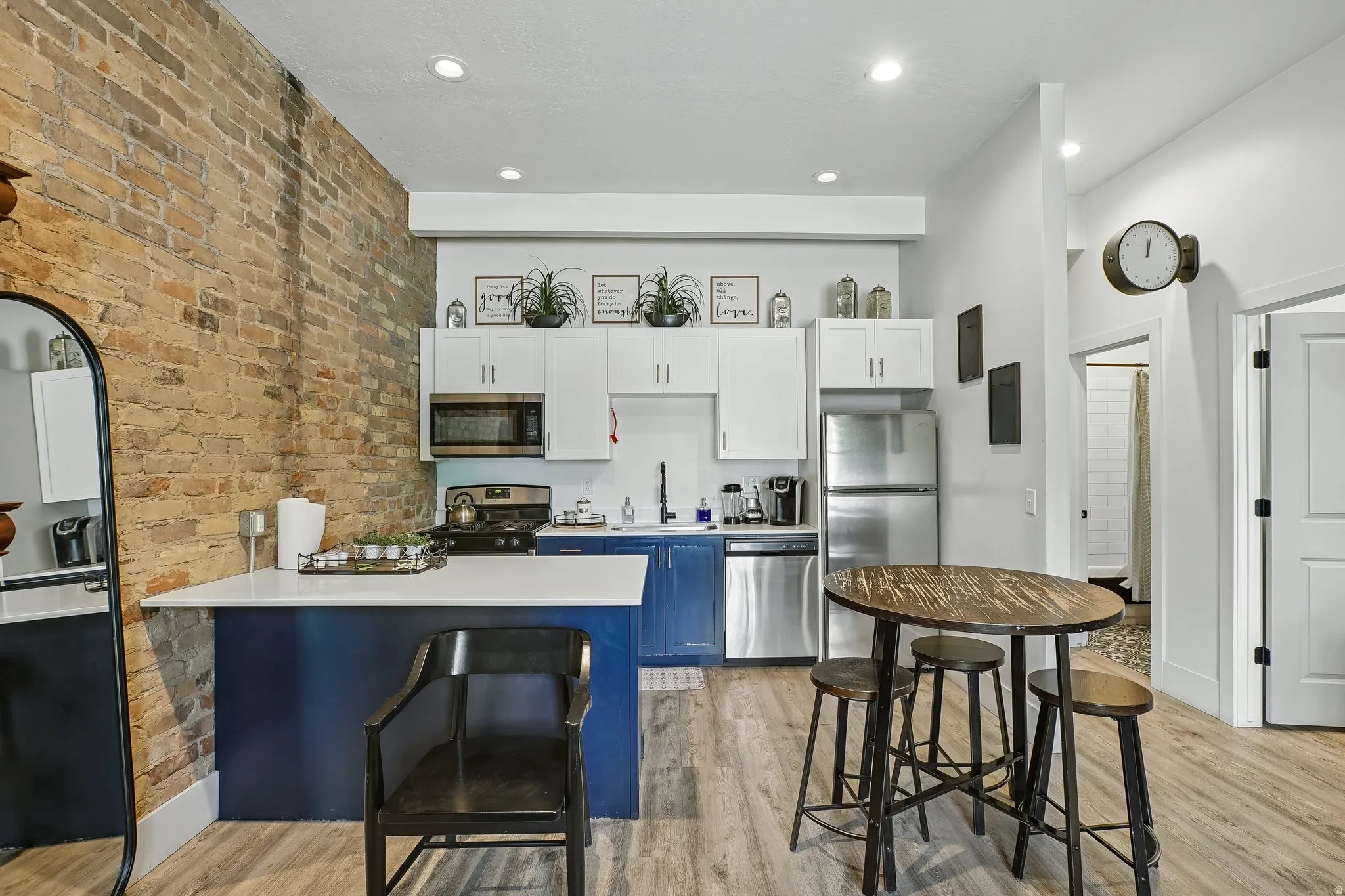 Modern kitchen and dining area featuring a blue-fronted island with waterfall countertop, stainless steel appliances including a refrigerator with glass doors, white cabinetry with integrated hardware, wood-finish flooring throughout, and a casual dining