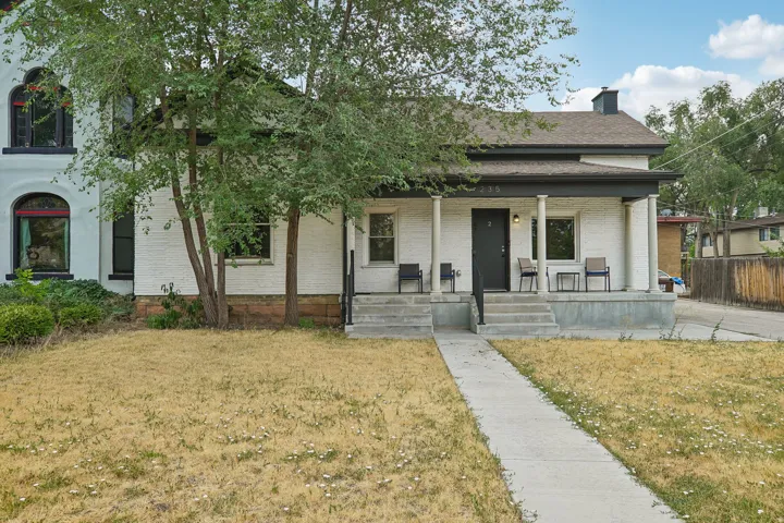 Classic front elevation featuring a white exterior with black trim, covered front porch with columns, mature shade trees, and a concrete walkway leading to the residence